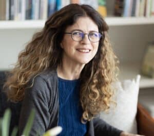 אישה מחייכת, בעלת שיער ארוך וגלי ומשקפיים, יושבת מול מדף ספרים.
