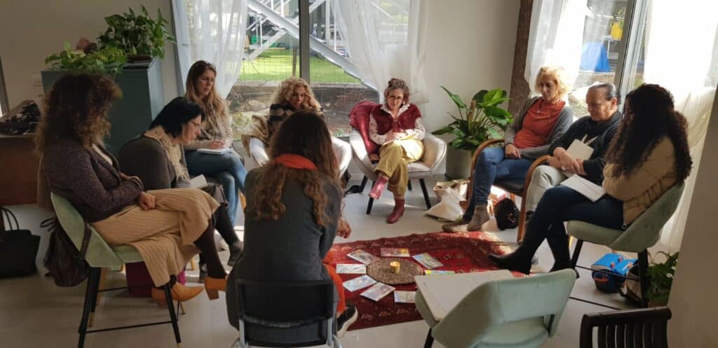 קבוצה של תשע נשים יושבת במעגל על כיסאות סביב שטיח קטן, ובמרכזו מונחים קלפים ונר.