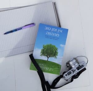 מחברת ספירלה, עט סגול, מצלמה דיגיטלית כסופה, וספר עם טקסט בעברית ותמונה של עץ על הכריכה, כולם מונחים על משטח לבן.