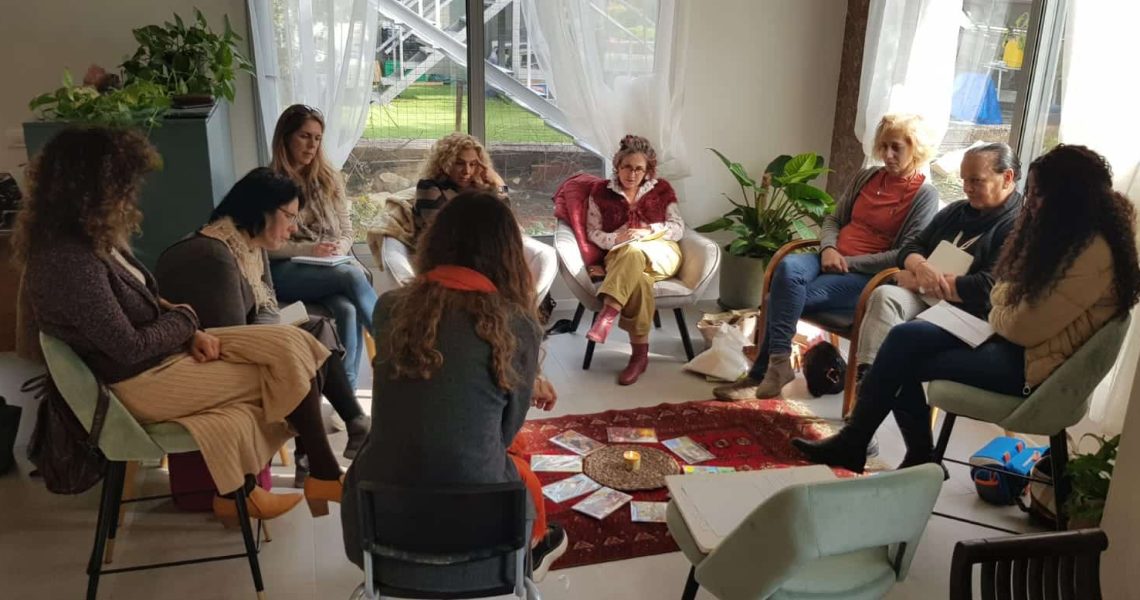 קבוצה של תשע נשים יושבת במעגל על כיסאות סביב שטיח קטן, ובמרכזו מונחים קלפים ונר.